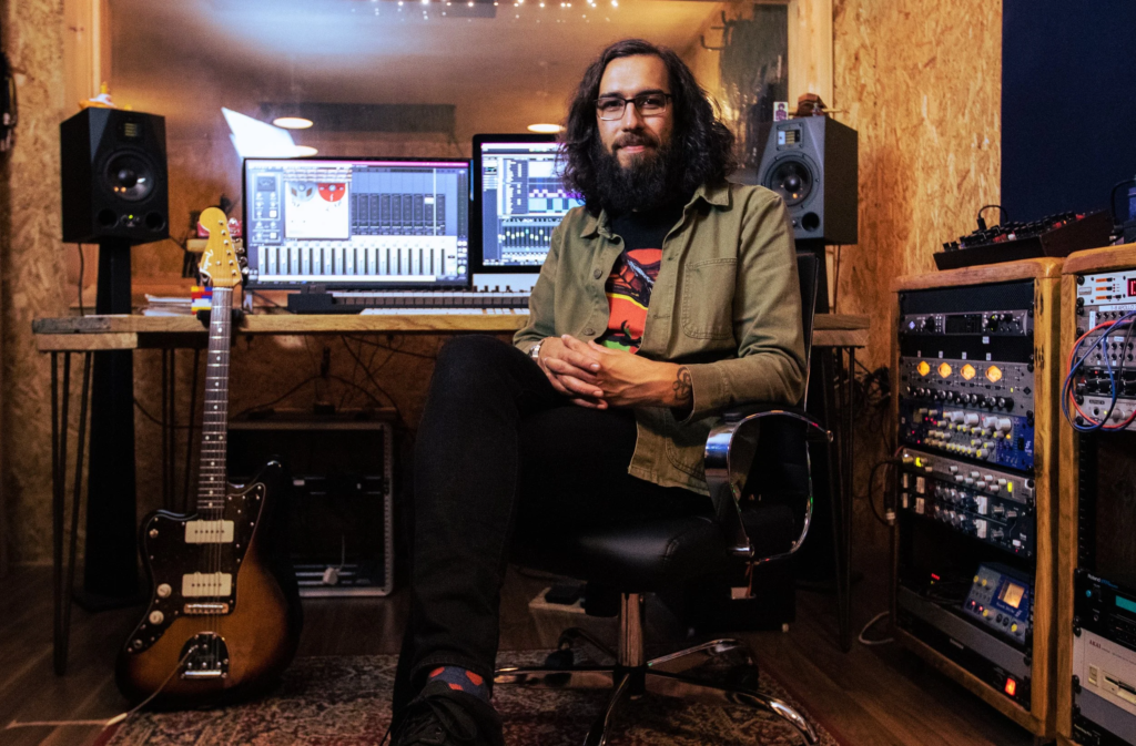 Jon Lawton sitting in a chair with the control desk for his recording studio behind me and an electric guitar. Photo by Lucy McLachlan